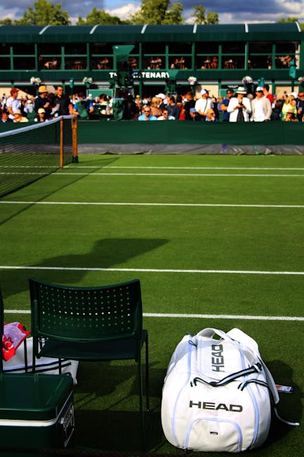 A view of a tennis court with vibrant green artificial grass marked by white lines, situated outdoors under clear weather. In the foreground, a black plastic chair and a matching small table are posit
