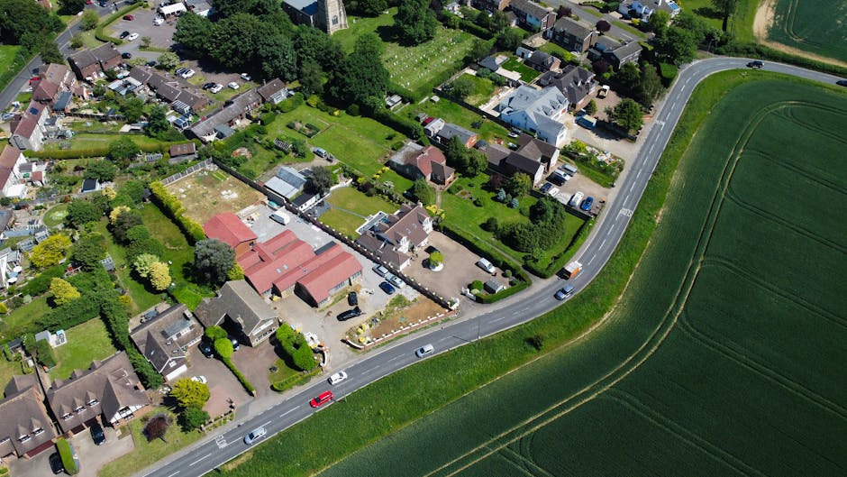 Aerial view of a residential area featuring multiple houses with varied roofing types, surrounded by well-maintained gardens, hedges, and trees. Some homes have driveways with parked cars, and visible
