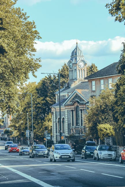 A city street scene with several cars parked along the side and others driving past. The street is lined with tall trees and historic buildings, including a prominent church with a clock tower topped